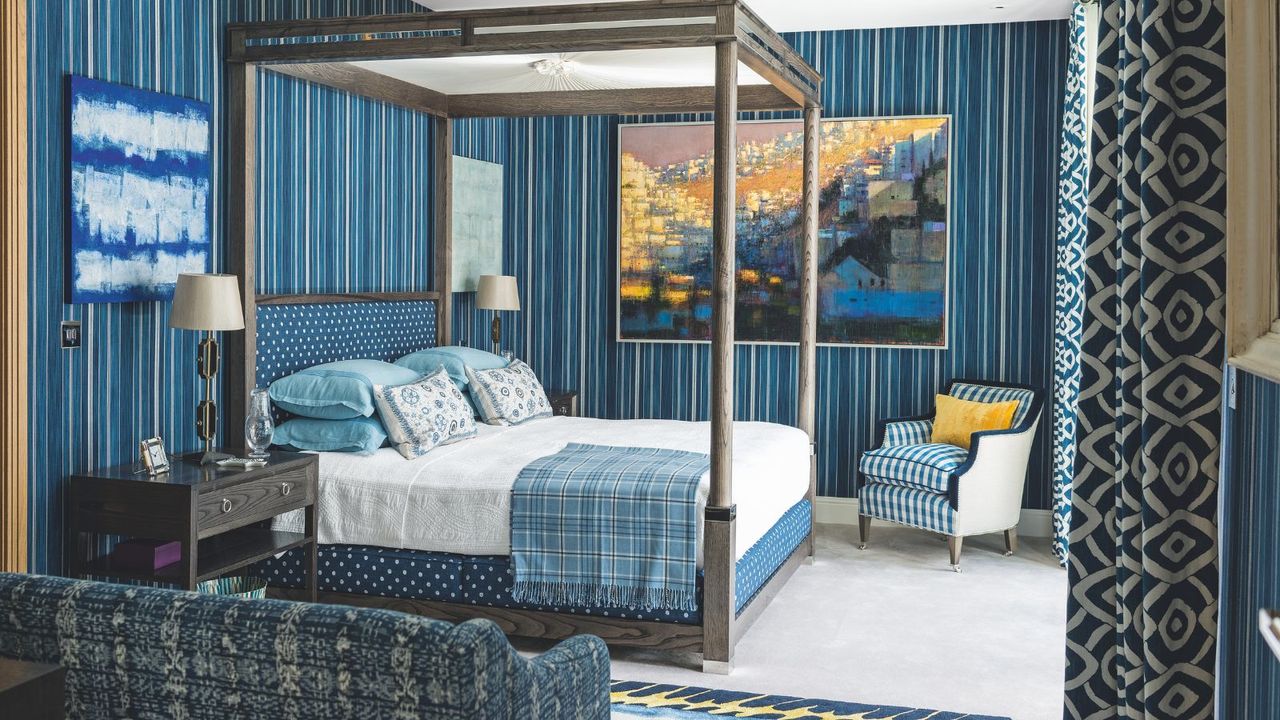A blue bedroom with striped blue wallpaper, patterned blue and white curtains, and white carpet. In the centre of the room is a large wooden four-poster bed, with white bedding, blue pillows and cushions, and a blue tartan blanket. At the foot of the bed is a blue and white striped upholstered chair with yellow cushion, and in the foreground of the image is a blue patterned couch. On the walls are various blue-themed artworks, and to the left of the bed is a wooden bedside table with a lamp with white lampshade on top.