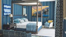 A blue bedroom with striped blue wallpaper, patterned blue and white curtains, and white carpet. In the centre of the room is a large wooden four-poster bed, with white bedding, blue pillows and cushions, and a blue tartan blanket. At the foot of the bed is a blue and white striped upholstered chair with yellow cushion, and in the foreground of the image is a blue patterned couch. On the walls are various blue-themed artworks, and to the left of the bed is a wooden bedside table with a lamp with white lampshade on top.