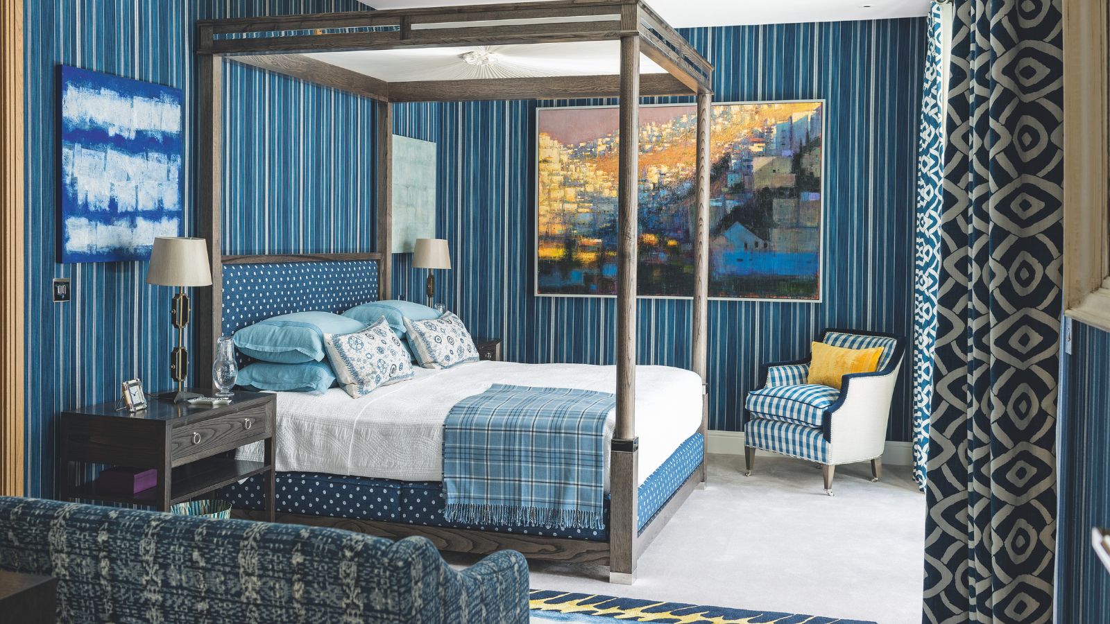 A blue bedroom with striped blue wallpaper, patterned blue and white curtains, and white carpet. In the centre of the room is a large wooden four-poster bed, with white bedding, blue pillows and cushions, and a blue tartan blanket. At the foot of the bed is a blue and white striped upholstered chair with yellow cushion, and in the foreground of the image is a blue patterned couch. On the walls are various blue-themed artworks, and to the left of the bed is a wooden bedside table with a lamp with white lampshade on top.