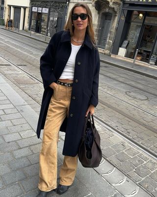 French fashion influencer @annelauremais wearing brown trousers, a white tank, navy coat, and black studded leather belt