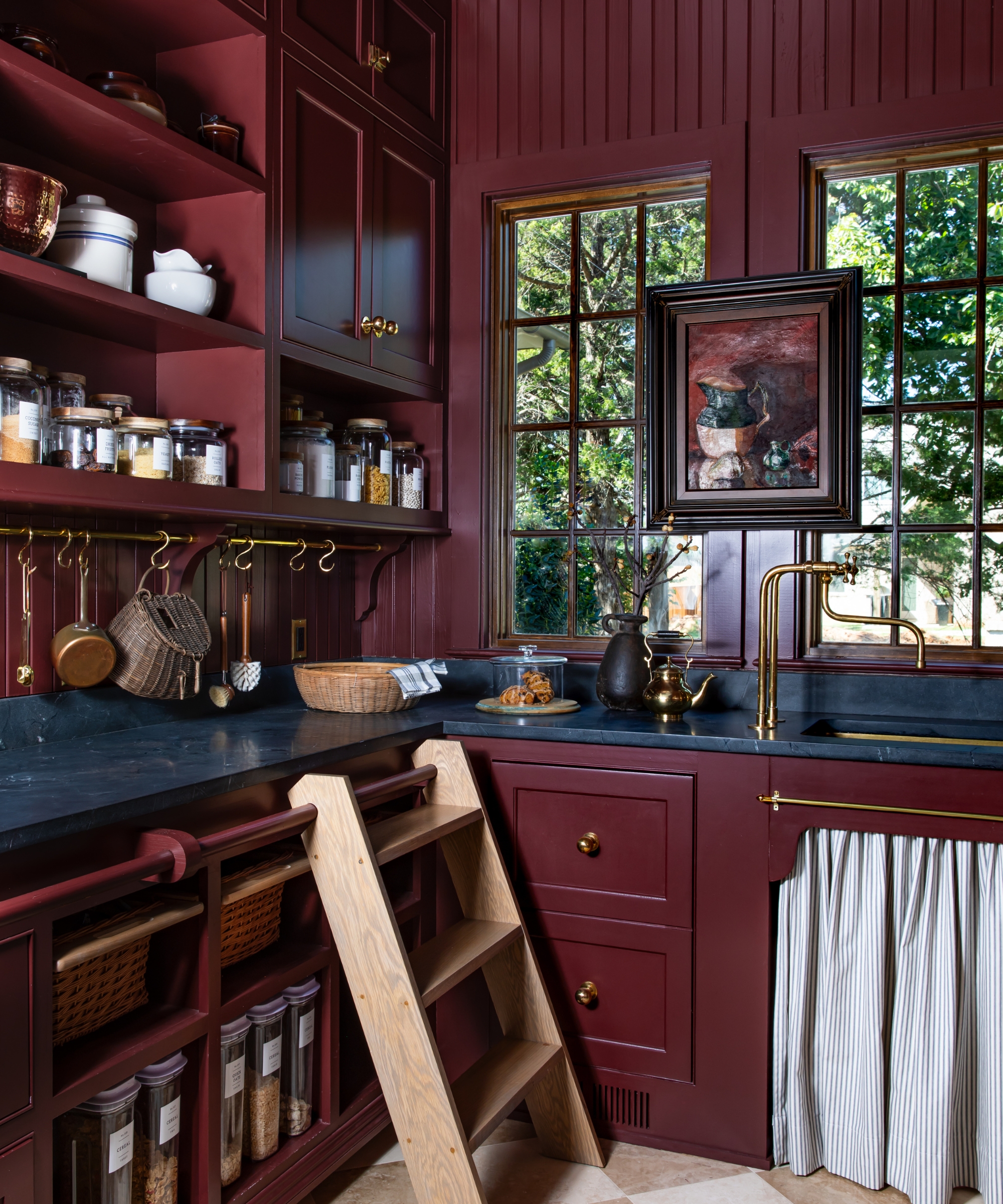 A moody red pantry with black countertops, open shelving on the walls, a wooden step ladder and a brass pot hanger on the wall
