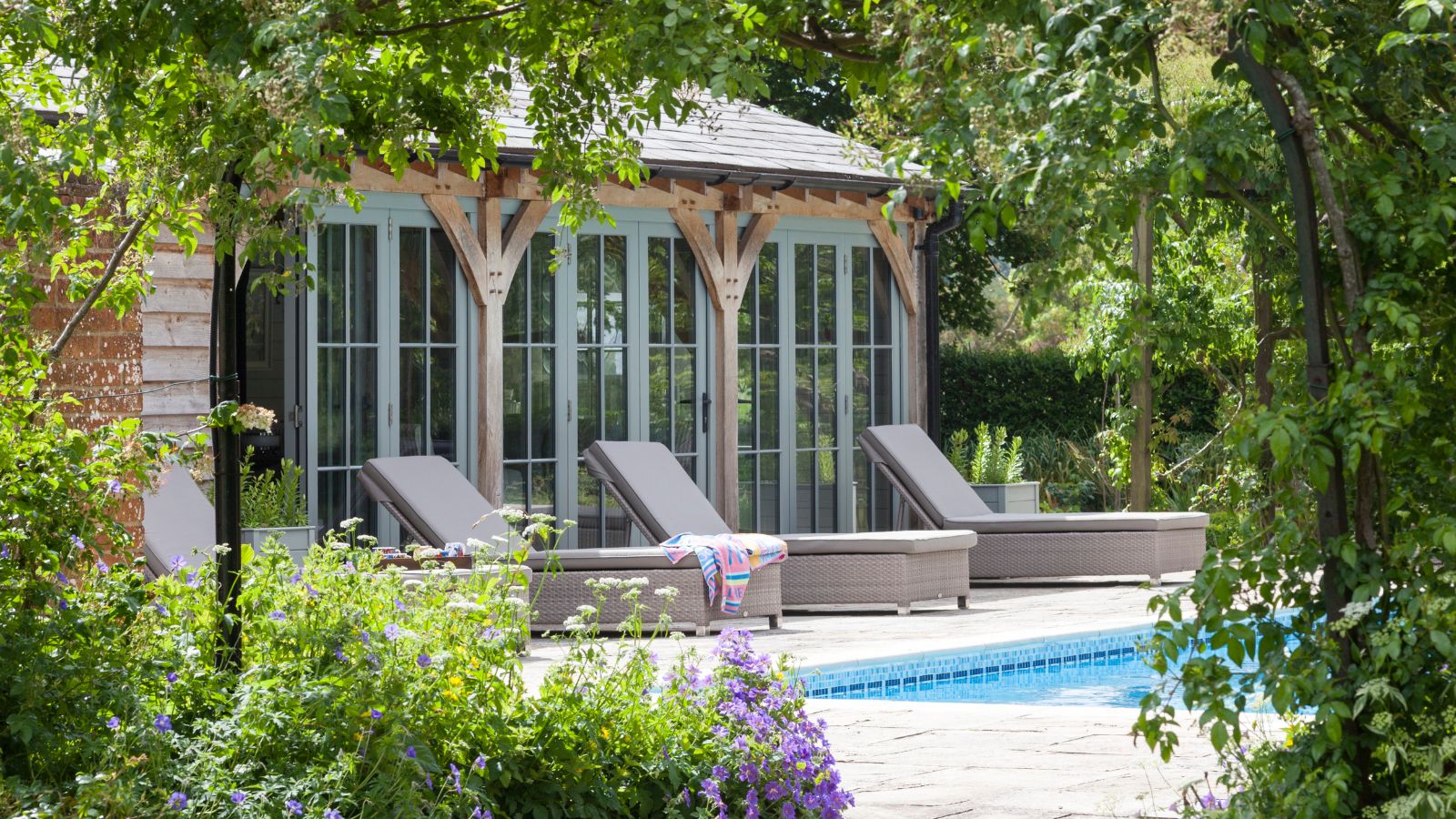 A leafy sunny garden with a large pool house, pool and loungers 