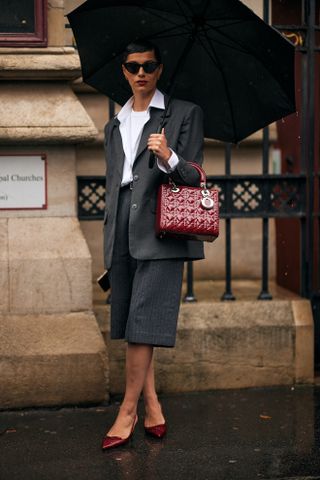 Woman at fashion week wearing preppy outfit featuring a white tee, white button down-shirt, gray blazer, gray shorts, and red heels.