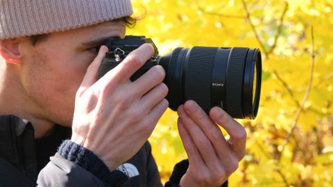 Sony a1 II camera with a Sony FE 28-70mm f/2 GM lens attached helf up to a person&#039;s face in front of some yellow trees