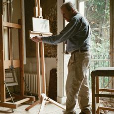 Lucien Freud painting at an easel whilst his whippet Pluto reclines on a leather chair