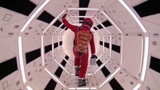 An astronaut in an orange space suit walks away from the camera while inside a space vessel, holding a small metal box