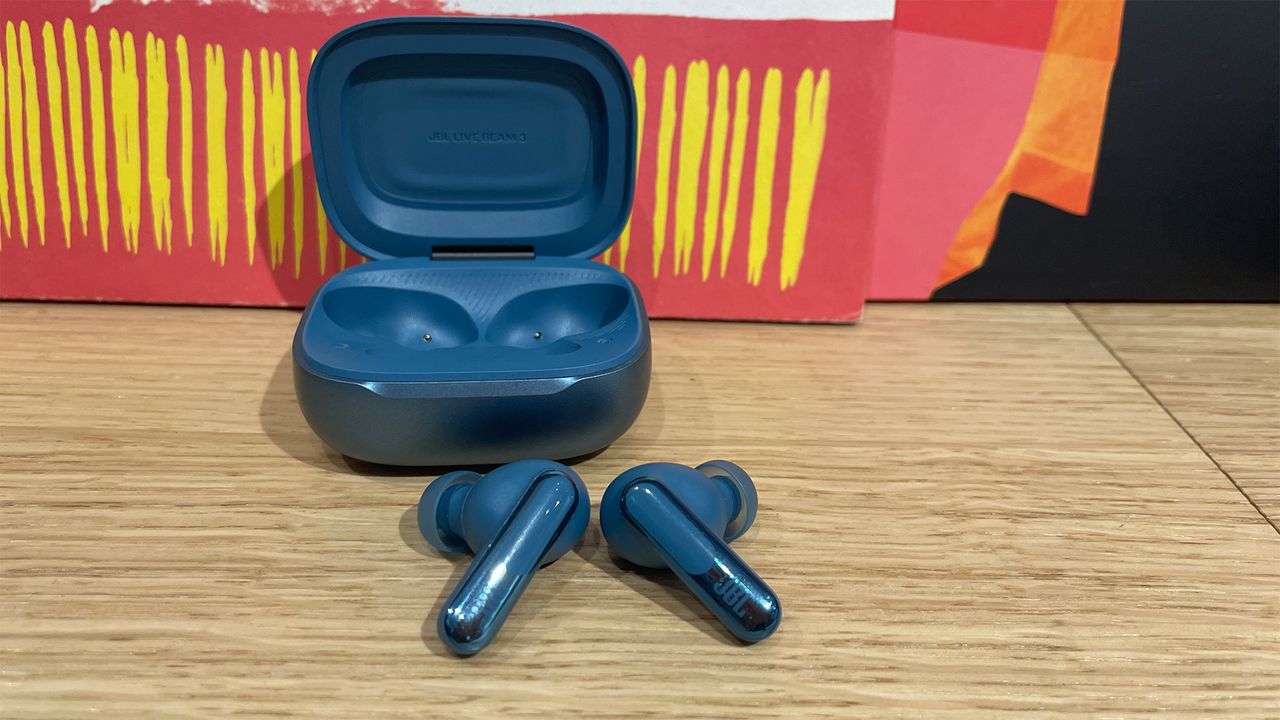 The blue case for a pair of JBL Live Beam 3 earbuds. The lid is open and both of the buds have been removed and placed on the wooden surface in front of the case.