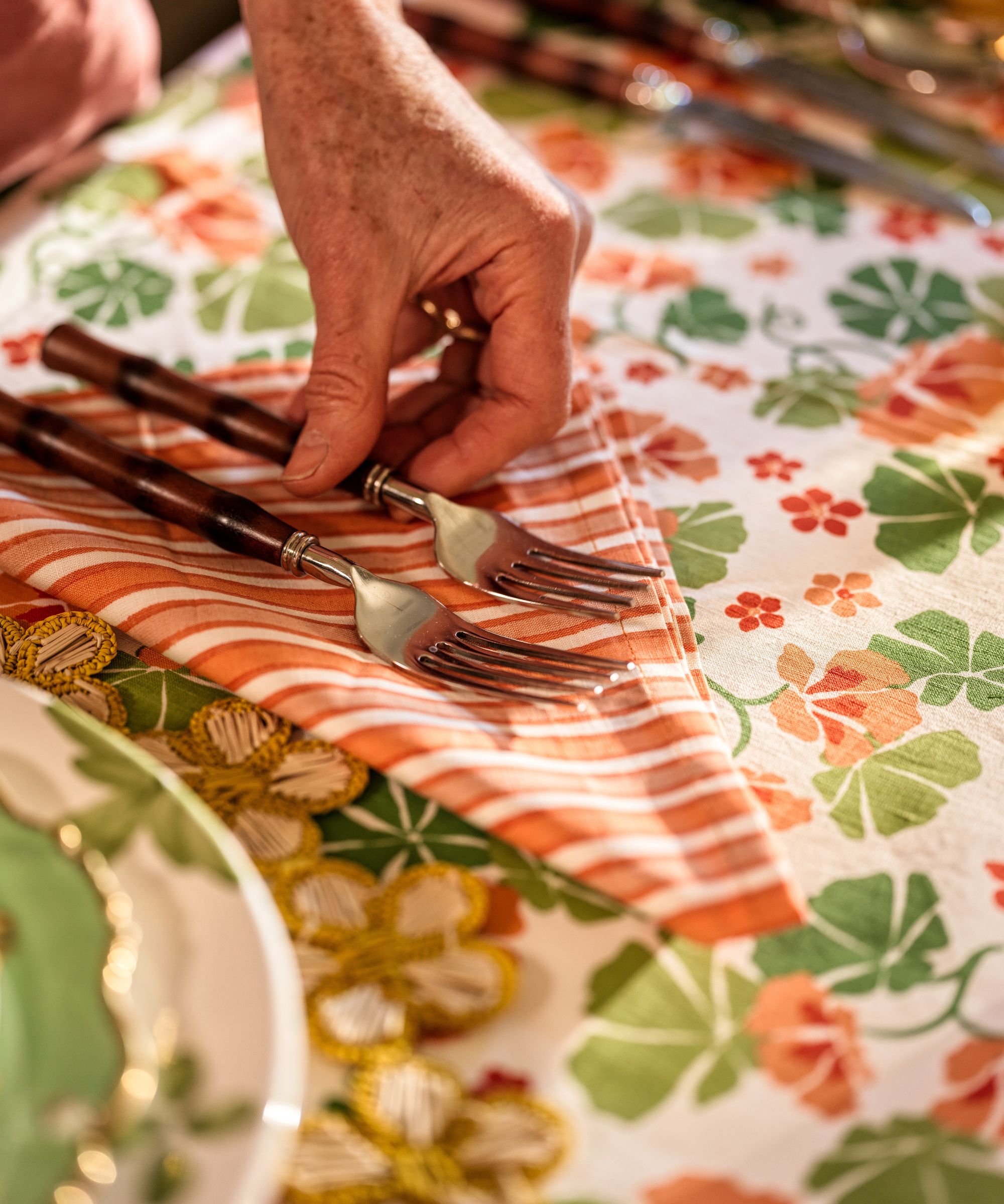 A close-up of a hand placing a pair of bamboo-handled forks onto an orange and white striped napkin, set against a green and orange floral tablecloth