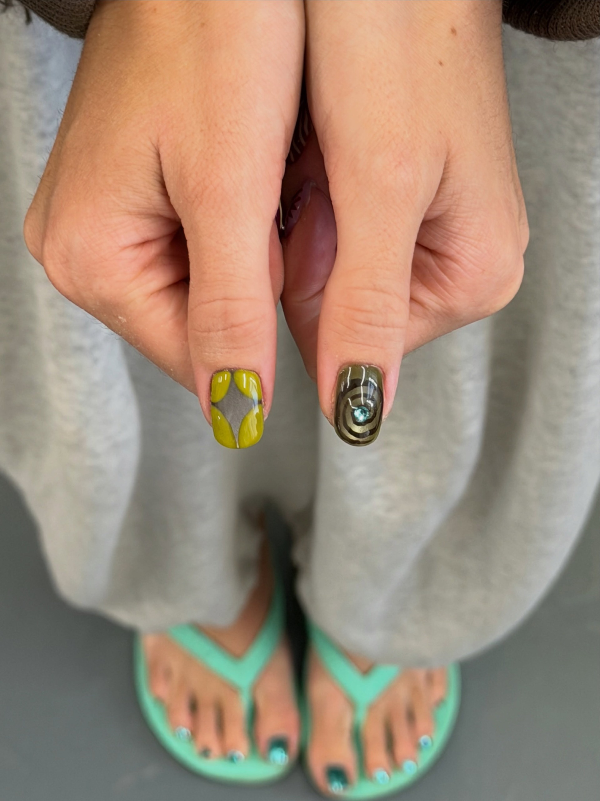 Two thumbs show colorful nail art, with one lime green and gray design and the other a metallic swirl pattern