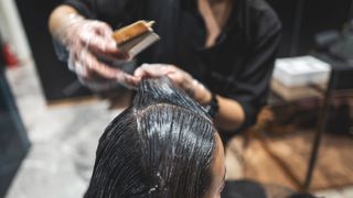 Close-up image of a hairdresser combing a white cream through a woman's long black hair in a salon. The woman's head in the foreground is in focus but the rest of the image is slightly blurred