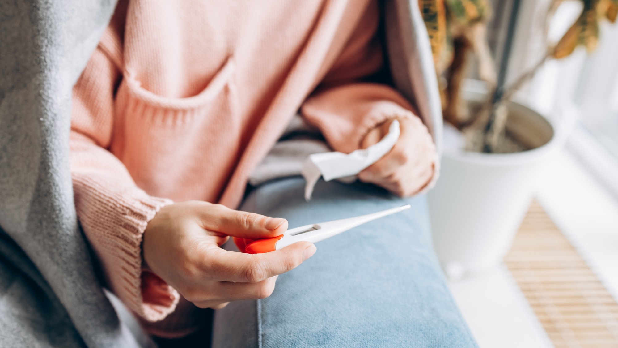 Close-up of woman holding thermometer and tissue while sitting under a blanket