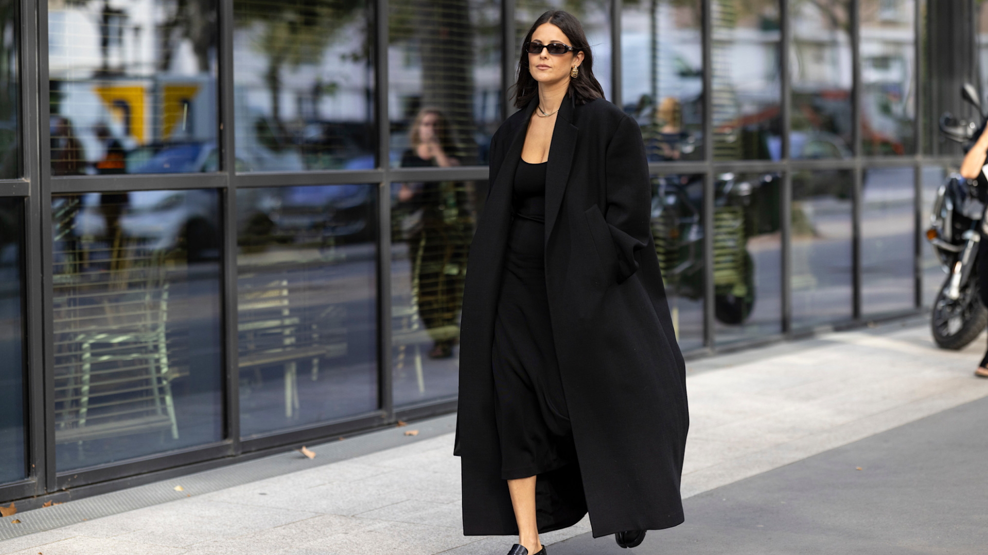 fashion week attendee wearing long black coat, sunglasses, black dress, and loafers
