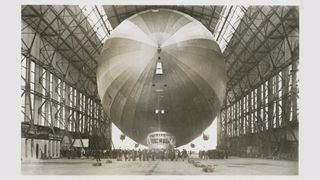 An old photograph showing the Graf Zeppelin inside a hanger. In front of it there are lots of people who look very small in comparison to the big airship.