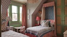 Bedroom with patterned wallpaper, a green window frame, a striped canopy single bed with blue-and-white bedding, a small desk beneath the window, and another single bed opposite with a floral cushion.