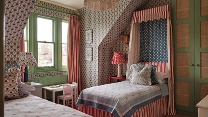 Bedroom with patterned wallpaper, a green window frame, a striped canopy single bed with blue-and-white bedding, a small desk beneath the window, and another single bed opposite with a floral cushion.