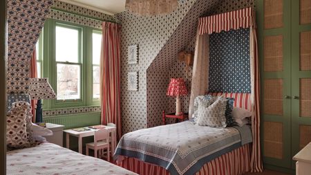 Bedroom with patterned wallpaper, a green window frame, a striped canopy single bed with blue-and-white bedding, a small desk beneath the window, and another single bed opposite with a floral cushion.