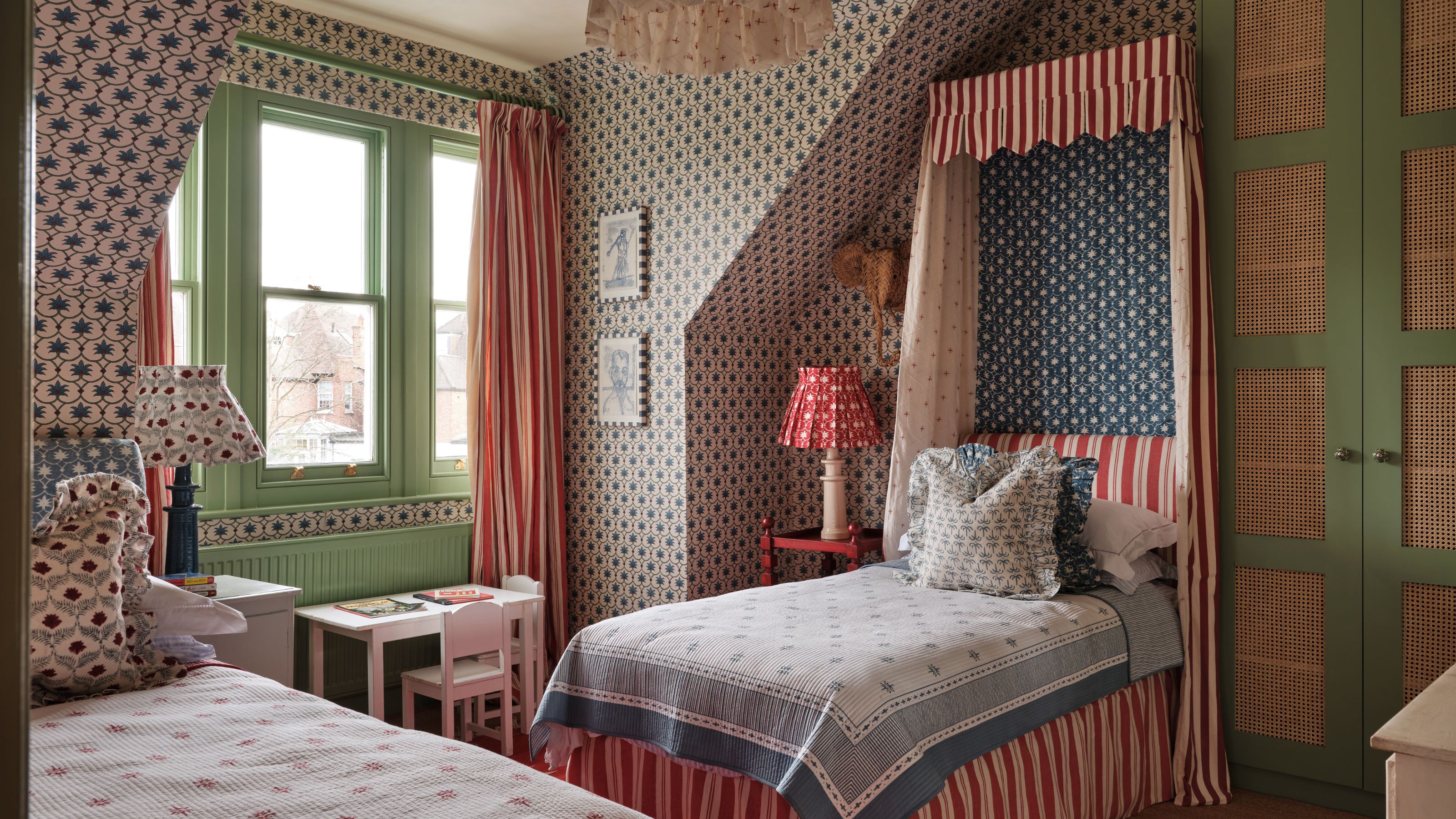 Bedroom with patterned wallpaper, a green window frame, a striped canopy single bed with blue-and-white bedding, a small desk beneath the window, and another single bed opposite with a floral cushion.