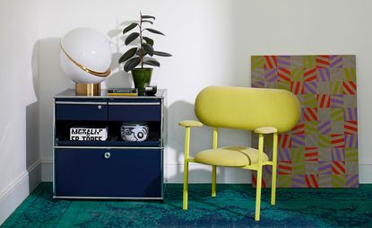 An interior vignette with a blue sideboard with spherical lamp on top, yellow armchair with oversized back and graphic artwork in the background, on the floor