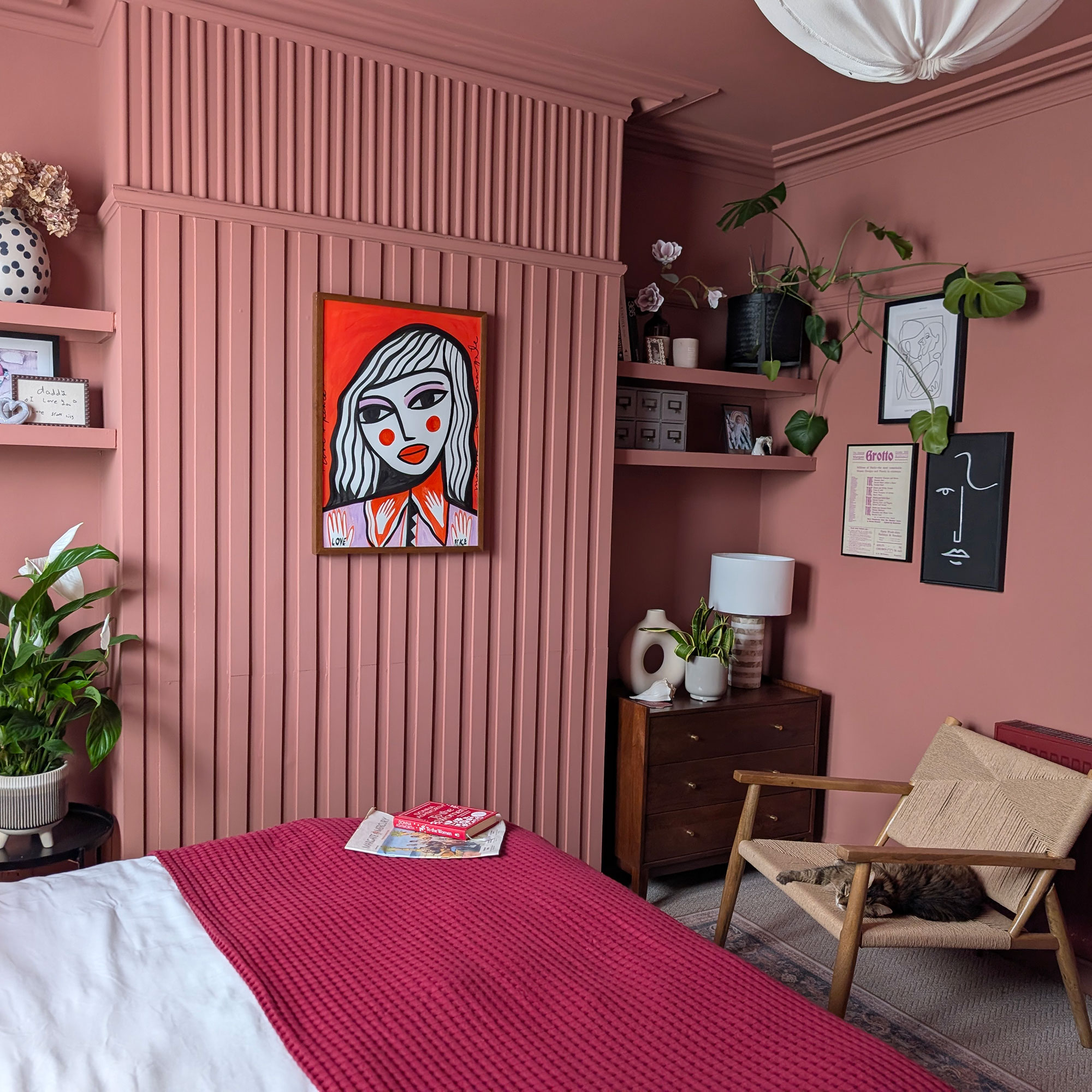 Pink bedroom with wall panelling, artwork of woman's face on wall, floating shelves and red throw on bed