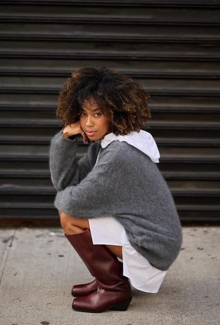 A woman crouching on the sidewalk wearing a white hooded shirt with a gray oversize sweater and brown knee-high boots.