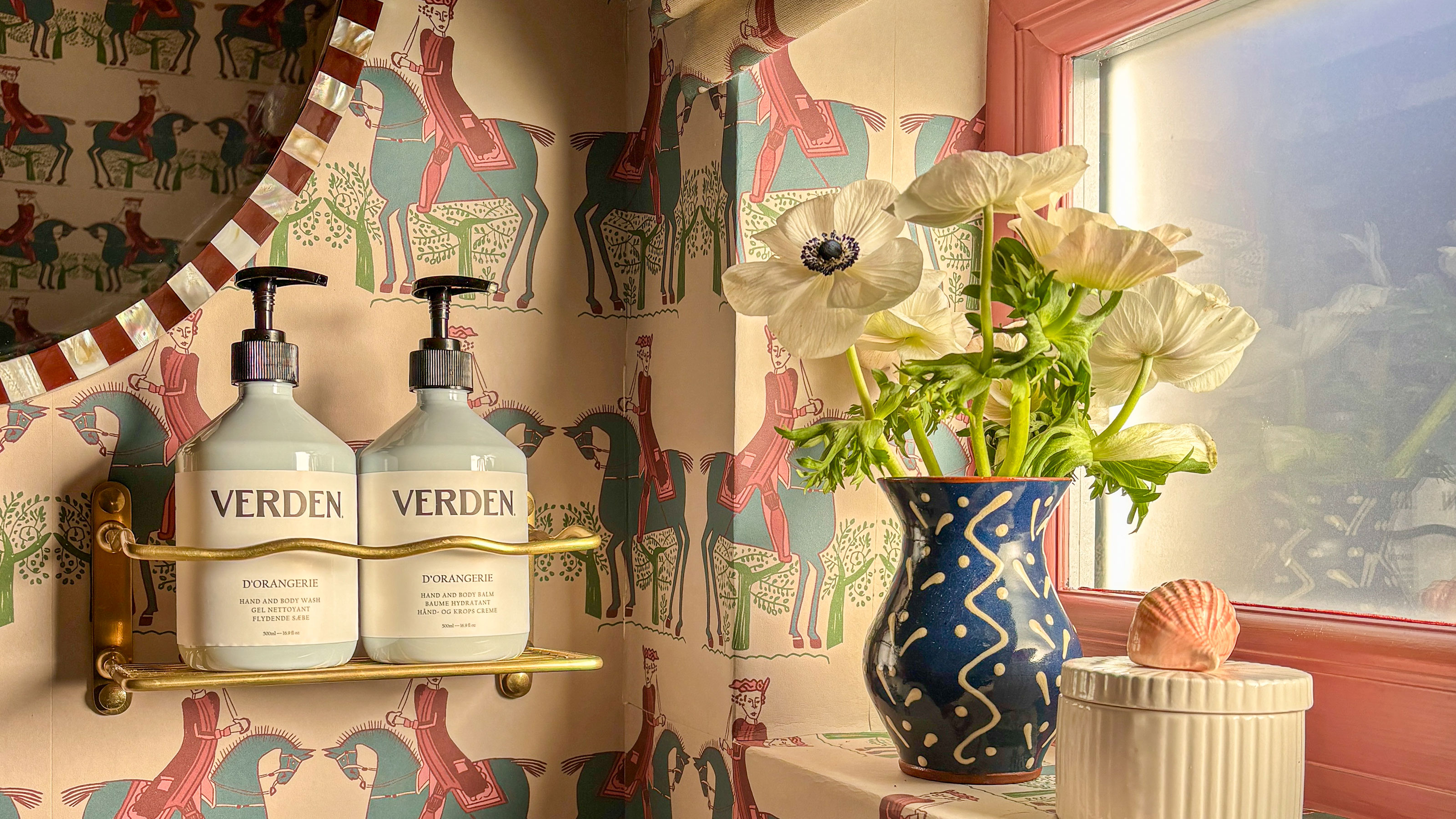 Corner of bathroom with horse patterned wallpaper, hand wash and lotion in gold brass holder and small vase of flowers on window sill