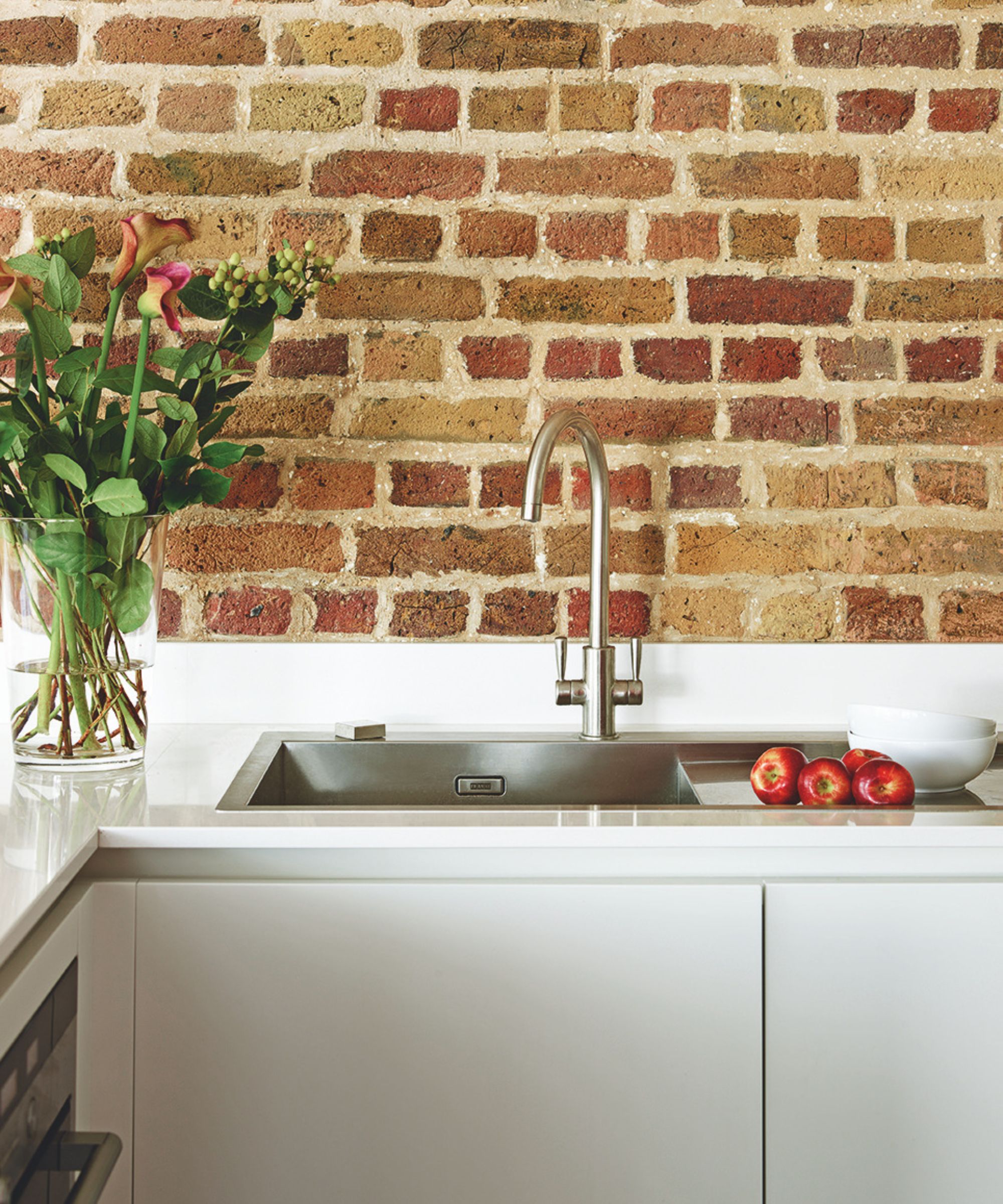 A stainless steel sink with silver faucet on a white countertop with large glass vase with pink flowers in front of an exposed brick wall.