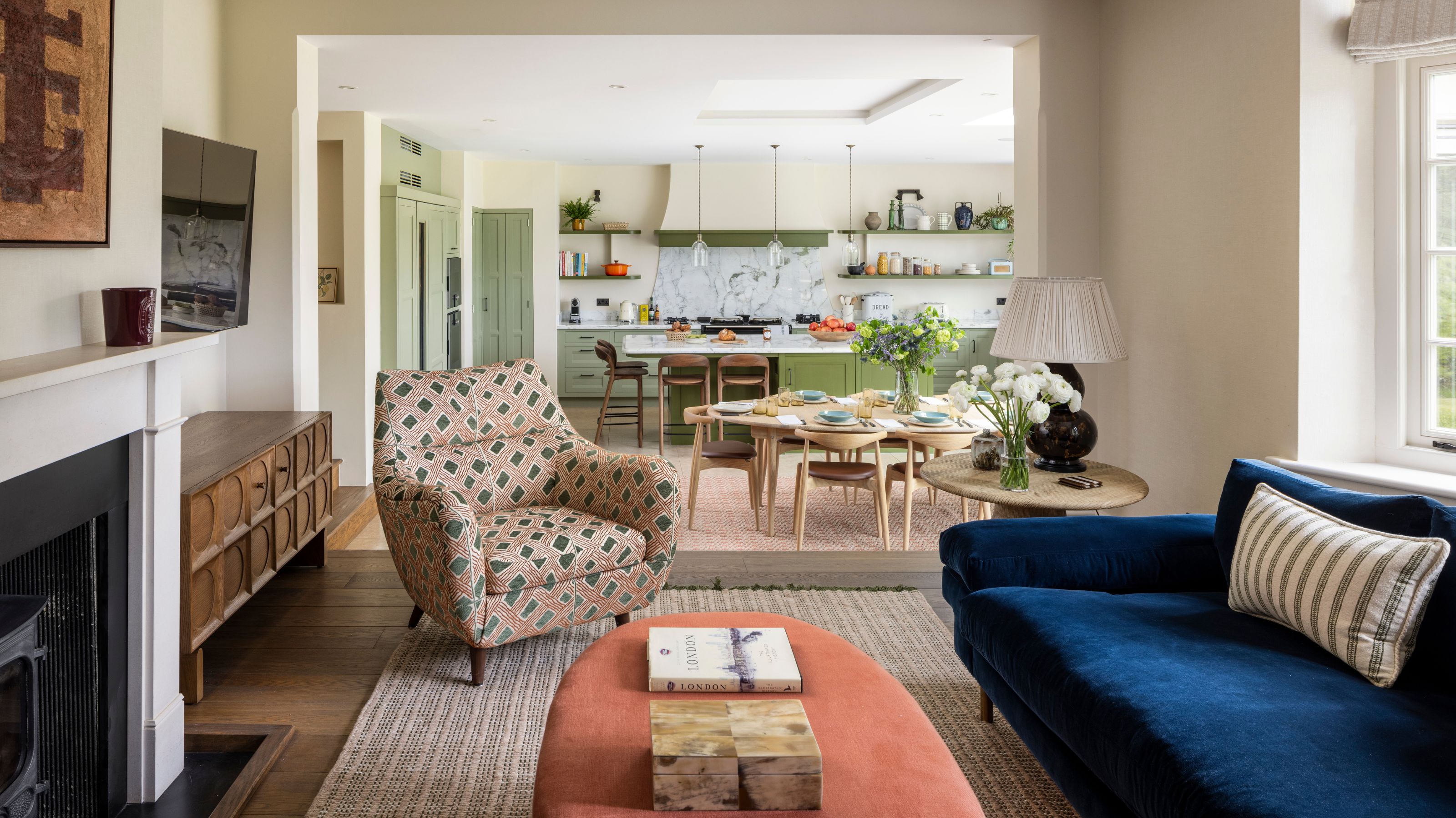a sitting area connected to a green kitchen with a fireplace, navy velvet couch, printed armchairs and vintage wooden furniture