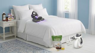 This image shows a mattress decorated in white linens in a calm, brightly lit blue room. On the bed is the FEPPO mattress vacuum while on the nightstand next to the bed is a bottle of DealMed Hydrogen Peroxide and a bottle of Cliganic Lavender Oil, plus a box of Ortho Bed Bug traps. In front of the bed is a Phueut steam cleaner and a bag of Harris Diatomaceous Earth