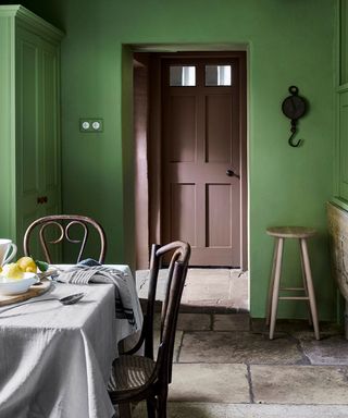Traditional dining and kitchen space painted in a vibrant green, traditional stone tile flooring, dining table with white tablecloth with two dark wooden chairs, light wood stool in background, green metal pendant light, looking through an open doorway to another room with brown painted door and walls