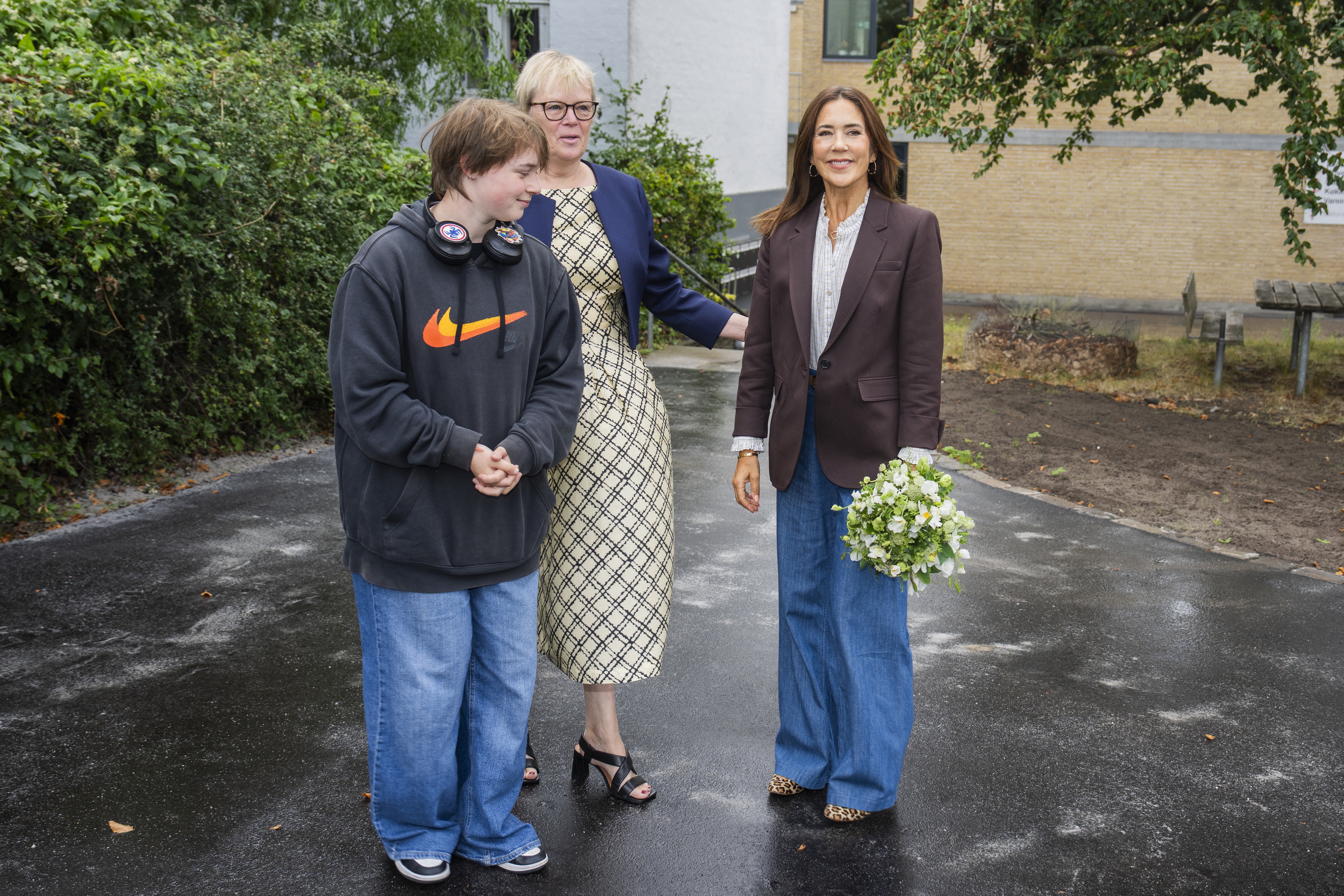 Queen Mary wearing jeans and a blazer standing with a teenage boy and woman