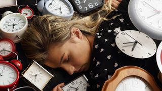 A woman with blonde hair sleeps surrounded by dozens of alarm clocks