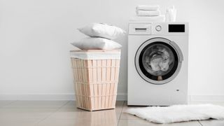 A washing machine running with two pillows stacked on a laundry hamper next to it
