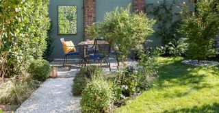 Backyard with evergreen planting, gravel path and green painted wall with garden mirror to show budget garden ideas