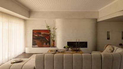 A minimalist living room with paneled ceilings, a firepit nook, a leaning art piece, a vase of stems, a ribbed greige couch, a coffee table with a vase of hydrangeas and a pair of candlesticks