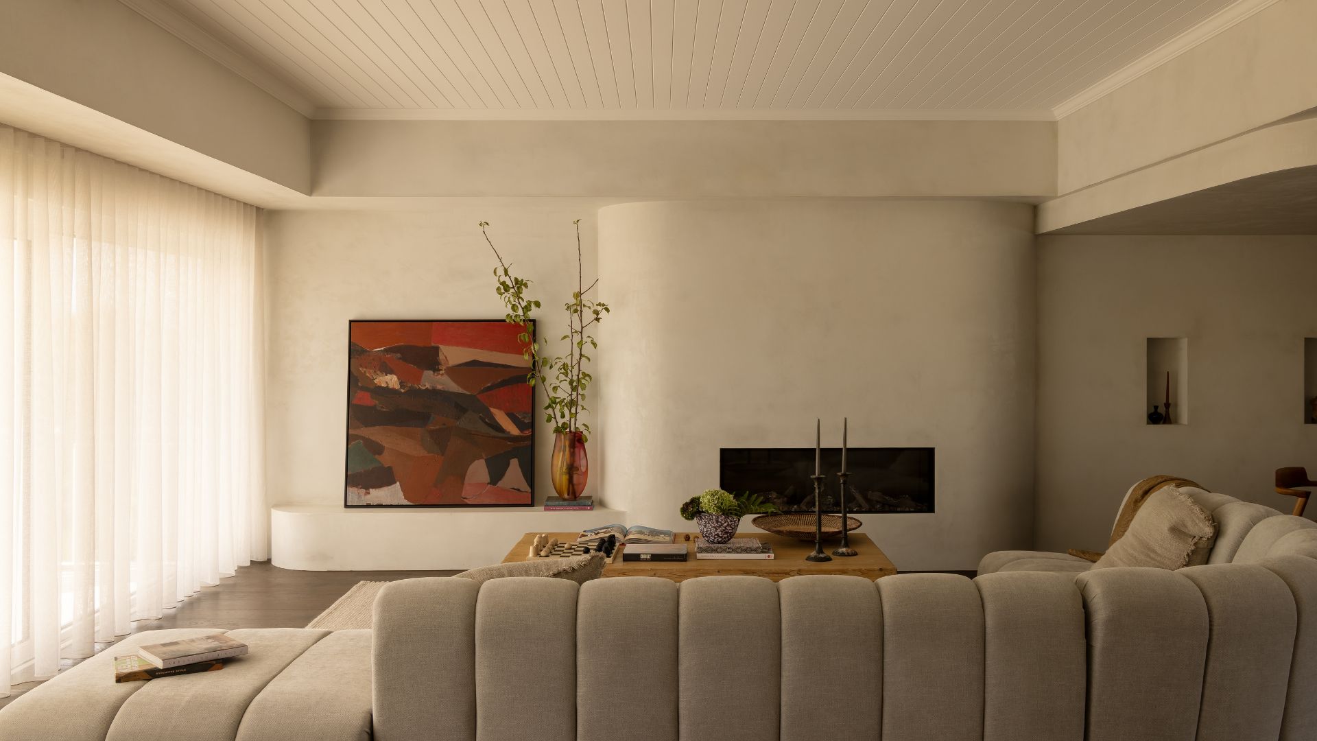 A minimalist living room with paneled ceilings, a firepit nook, a leaning art piece, a vase of stems, a ribbed greige couch, a coffee table with a vase of hydrangeas and a pair of candlesticks