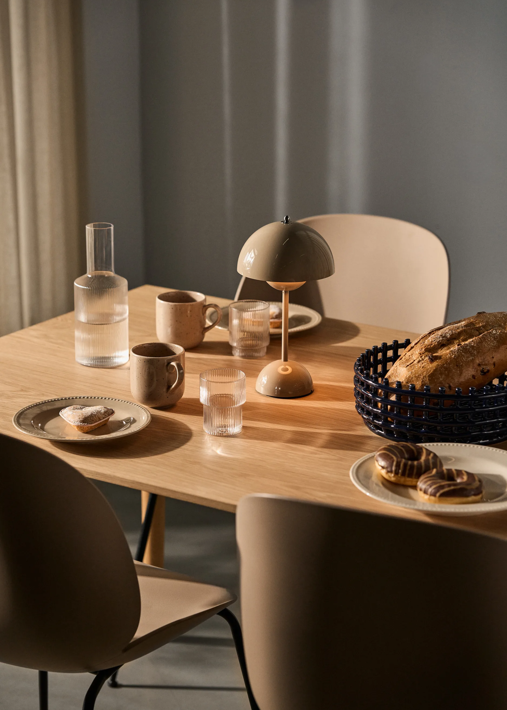 A table set with plates of pastries, coffee mugs, glasses, and a bread basket beside a lamp