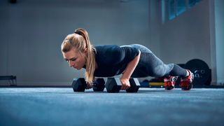 Woman in gym performs pushup holding dumbbells