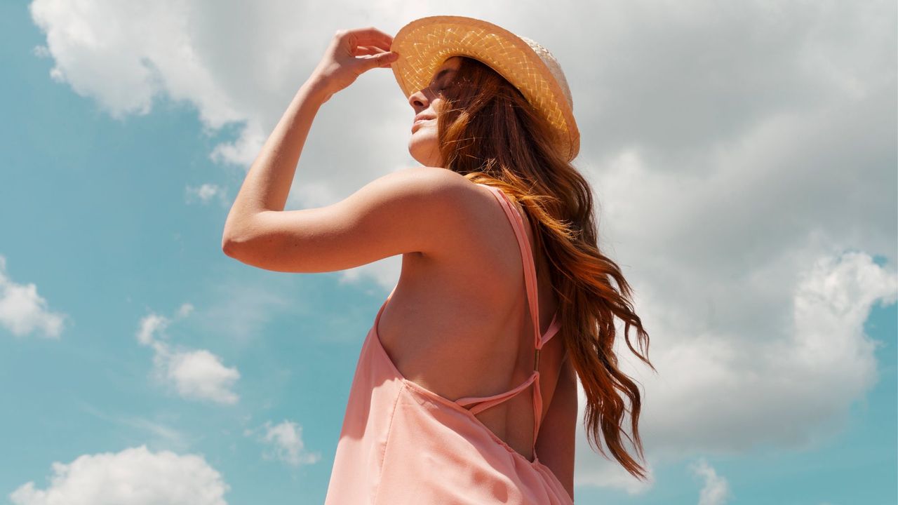 A woman with long, red hair wearing a pink strappy sun dress and a straw hat looking up to the sun in the sky