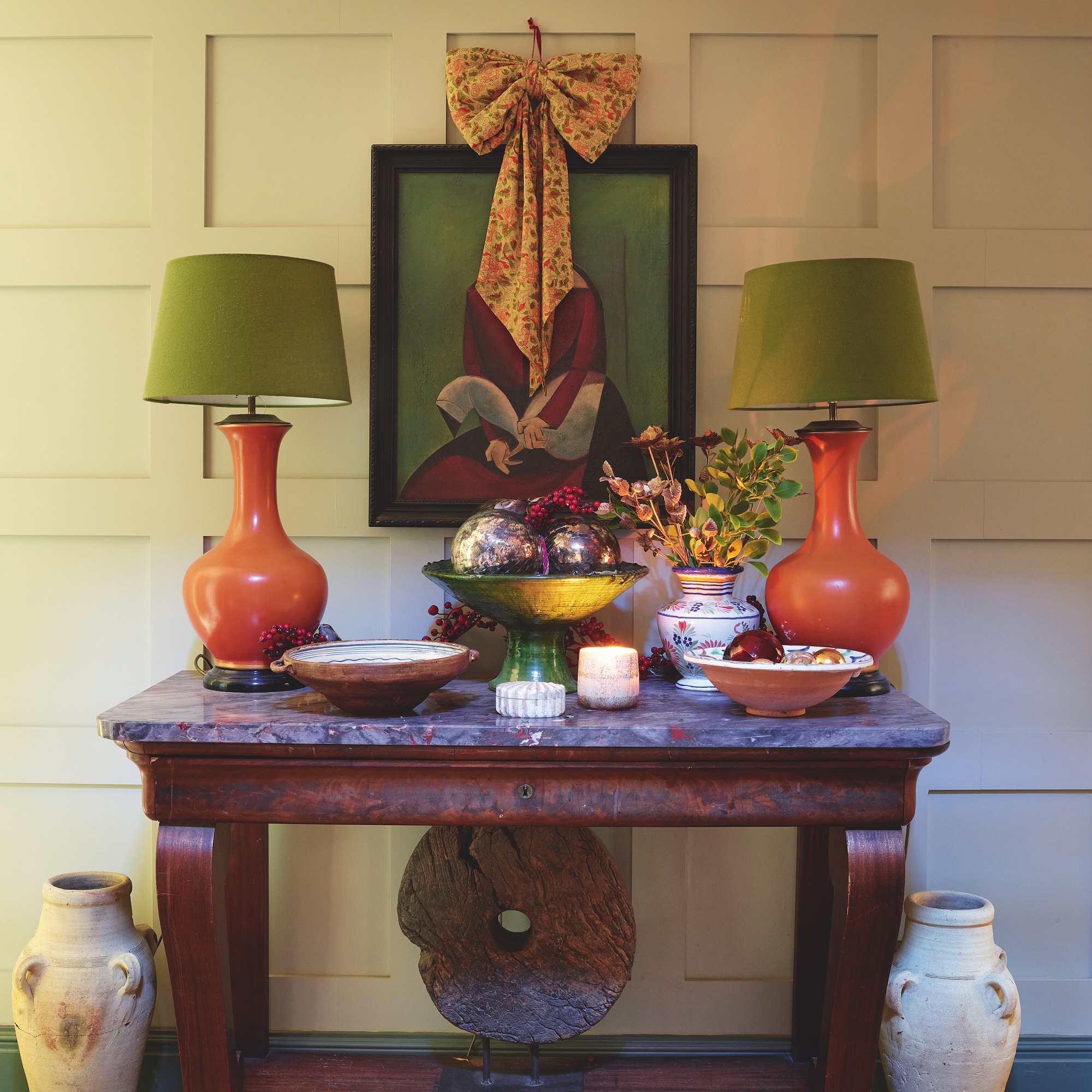 antique marble top console table with two orange lamps with green shades and artwork above decorated with bow