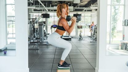 Fit female athlete performing squats in a gym with dumbbells in hand