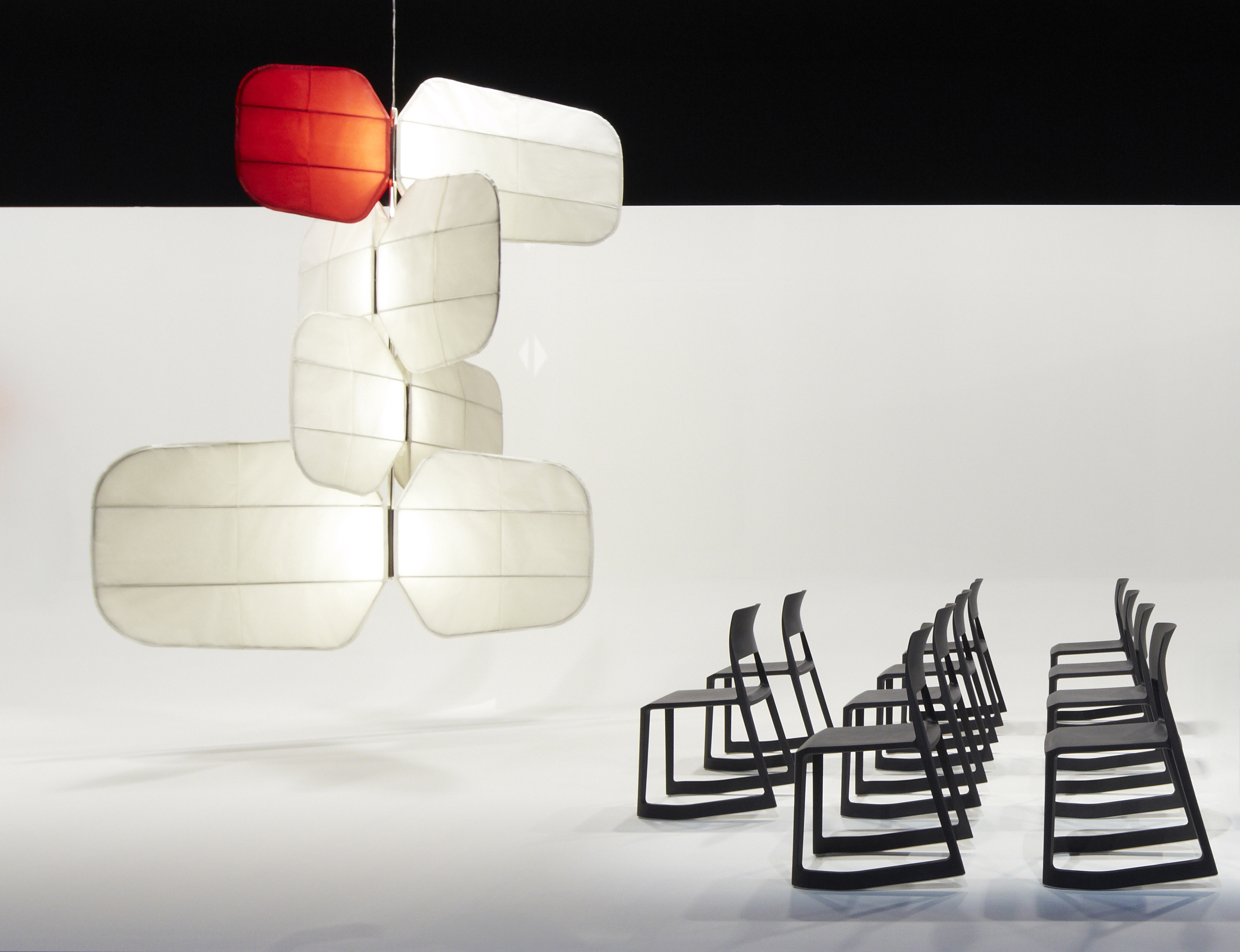 A design installation featuring a series of rocking black chairs with a minimalist silhouette and a pendant white and red ceiling lamp made with multiple abstract, petal-like components.