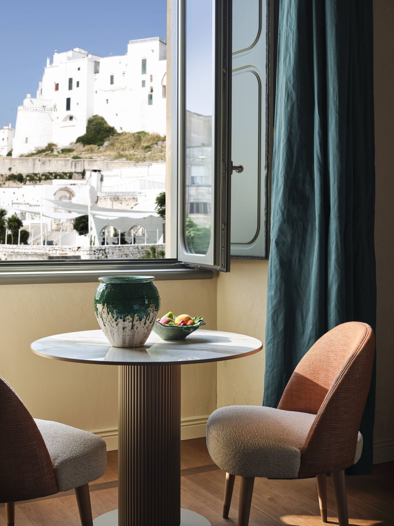 A beautiful, sun-lit view of a white-washed old town can be seen from the inside of a hotel room furnished with sculptural pieces like a round table, upholstered chairs in coral textiles, and wavy curtains.