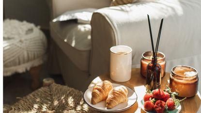A reed diffuser on a side table with candles and croissants next to an arm chair