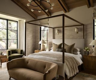 Wood beamed bedroom with cream walls, neutral bedding, brown sofa, green armchair and large windows