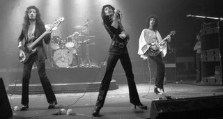 A black-and-white shot of Queen performing live in 1974, with John Deacon on the left, Freddie Mercury in the middle and Brian May wearing a white shirt on the right. Roger Taylor is in the background.