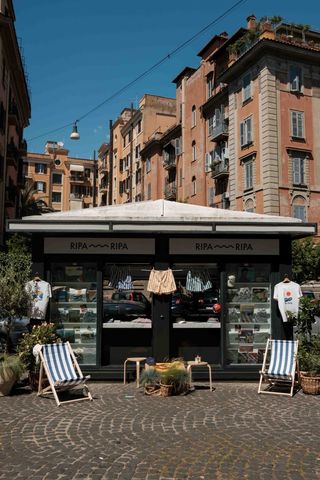 The outdoors of a newsstand features blue and white deck chairs, wooden tables, and swimming trunks, standing against brick-shaded tall buildings.