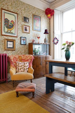 A classic living room with printed walllpaper, upholstered orange armchair, wooden drinks cabinet, wooden dining table and bench, with vase and floral bouquet