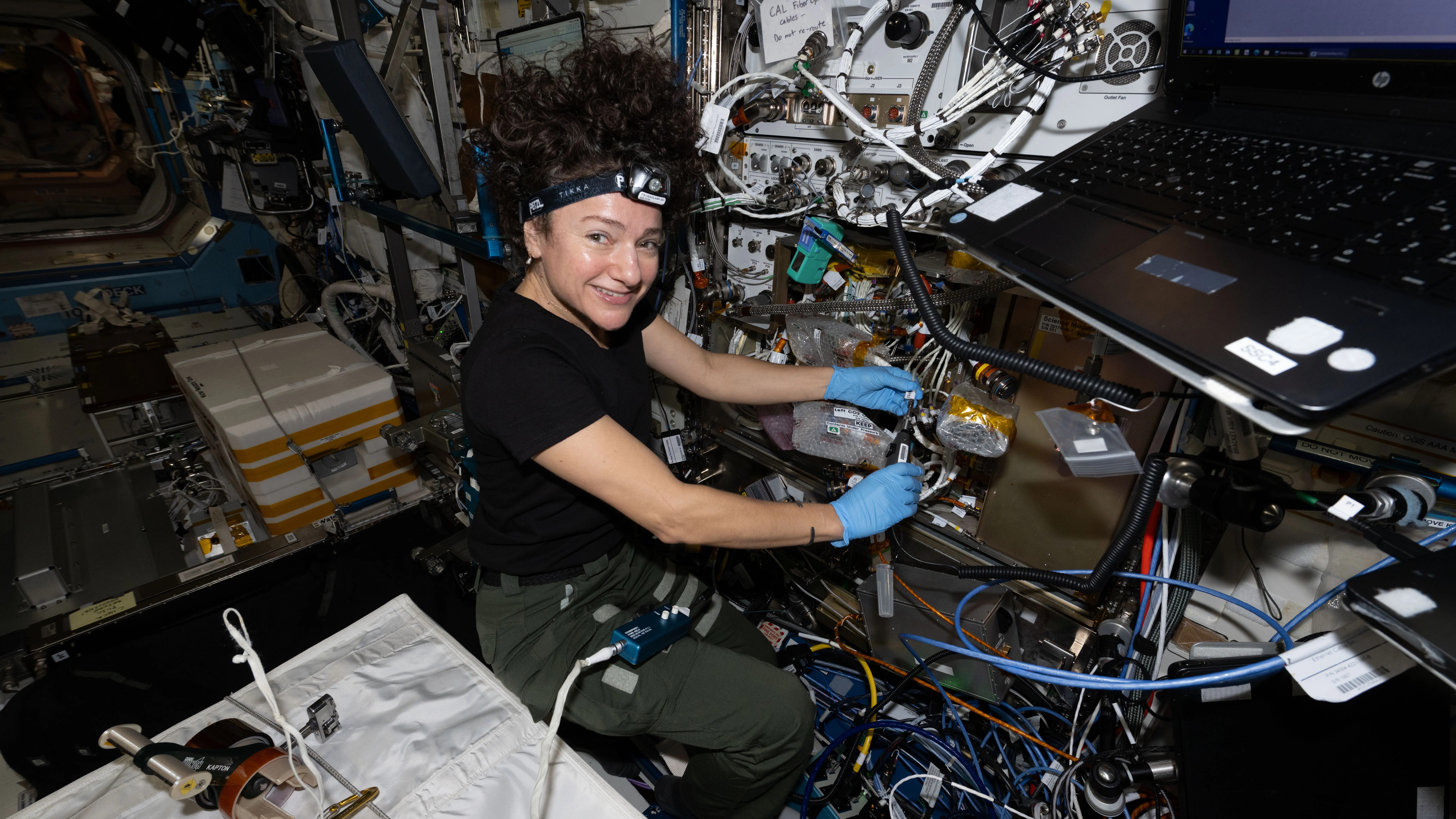 A woman in zero-gravity looks to the camera as she fixes equipment.