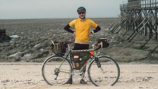 A white man stands behind his touring bike in a yellow jersey and gilet