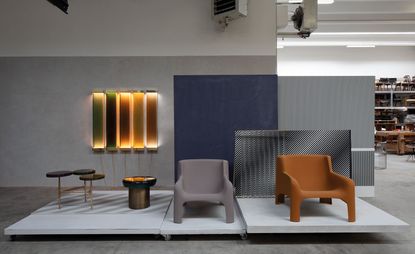 Two Contemporary side tables and two armchairs (grey and orange), displayed on a grey platform with colourful wall lights on a grey wall 